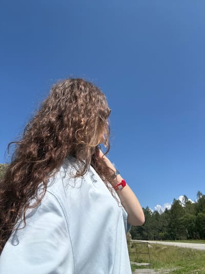 The image shows a person with long, curly brown hair facing away from the camera and looking towards a clear blue sky. They are wearing a light blue t-shirt and have a red wristband and a watch on their left arm. In the background, there is a forest with green trees, a dirt road, and a cloudy sky. The scene appears to be in a natural outdoor setting, possibly near Bichiktu-Boom, Russia, given the vegetation and the presence of a road.