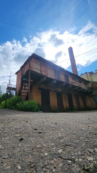 An exterior, ground-level view features a three-story brick building with metal supports and stairs, a chimney stack reaching skyward, alongside a cracked asphalt road under a cloudy sky. The building has evenly spaced, dark-colored doors or openings along the facade, with vegetation growing near its base. The scene suggests an industrial area or historical structure, potentially in a state of disuse. The location is Smolensk, Russia.