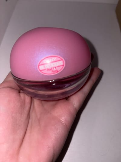 A pink, spherical perfume bottle labeled "DKNY BE DELICIOUS" is held in a person's palm. The bottle has a clear glass base and a pink, pearlescent cap. The background is a plain, light-colored surface. This image was taken in the city of Zasеchnoye, Russia.