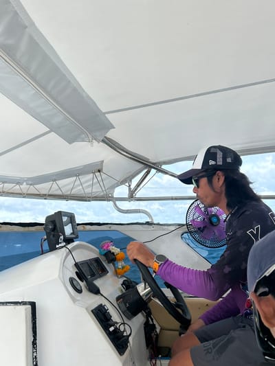 In Ratsada, Thailand, a person with long dark hair and a black and white baseball cap is operating a boat. They are wearing sunglasses and a black long-sleeved shirt with purple sleeves underneath. The person's hands are on the steering wheel, and they have an orange smartwatch on their left wrist. In the background, a purple fan is attached to the boat's interior. The boat has a white dashboard with various electronic devices, including a fish finder, and a decorative floral garland is hanging from a fixture. The boat is on the water, with a cloudy sky visible in the distance. Another person, wearing a blue hood and sunglasses, is partially visible in the foreground, seated to the right of the operator. The scene takes place under a white canopy that provides shade.