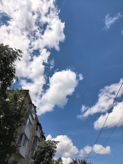 This media file is a photograph taken from a low angle, looking up at a bright blue sky filled with fluffy white cumulus clouds. A portion of a multi-story building, likely an apartment building, is visible on the left side of the frame. The building has a pale yellow facade and features several balconies with white railings. Dense green trees partially obscure the building on the left and right. Electrical wires crisscross the sky, with one prominent dark wire running diagonally from the top right towards the bottom center of the image. A street light is visible near the bottom of the frame. The scene suggests a daytime setting, likely late morning or early afternoon, given the bright sunlight illuminating the clouds and the clear blue sky. The weather appears to be fair and pleasant, with scattered clouds indicating a typical summer day. The location is identified as Bryansk, Russia. There is no visible text in the image. The overall mood conveyed is one of a serene, everyday urban landscape under a beautiful sky.