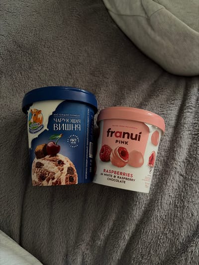 Two containers of frozen treats are positioned side-by-side on a textured, dark gray blanket, possibly a bedspread or throw, in a location identified as the city of Новинки, Russia. The container on the left is predominantly blue and white, labeled "КОРОВКА из Кореновки" (KOROVKA from Korenovka) and features the flavor "ЧАРУЮЩАЯ ВИШНЯ" (CHARMING CHERRY) with an image of cherry-swirled ice cream, a fresh cherry, and chocolate brownie pieces. Text indicates it is "НАСТОЯЩИЙ ПЛОМБИР" (REAL PLOMBIR) and adheres to "ГОСТ 31457-2012". The container on the right is pink and white, branded "franui PINK," and advertises "RASPBERRIES IN WHITE & RASPBERRY CHOCOLATE," displaying images of whole raspberries and raspberry-filled chocolates. It states "RECIPE FROM PATAGONIA" and "GLUTEN-FREE," with a net weight of 150g. A faint, lighter fabric is visible in the upper right and lower left background, suggesting additional soft furnishings.