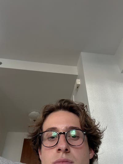 The image provides a low-angle, close-up indoor view, primarily featuring the lower half of a person's face. The individual has wavy brown hair, brown eyes, and wears round, dark-framed glasses, which exhibit reflections of a grid-patterned source. Visible facial features include the nose and upper lip. The background consists of light-colored walls and a flat ceiling with clean architectural lines. A recessed light fixture is partially visible on the upper left, above the outline of a dark wooden door. A small, white, rectangular device is mounted high on a wall or pillar to the right. No identifiable landmarks or exterior views are present to determine a specific location within Boston, United States.
