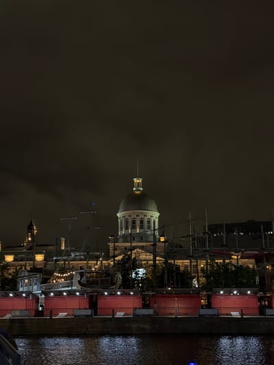 This image captures a nighttime scene of the Old Port of Montreal, Canada. The central focus is the illuminated Bonsecours Market, a large, light-colored building featuring a prominent dark dome topped with a light-emitting spire. To the left of the market, a smaller, darker building with a pointed spire is partially visible, also lit.

The sky above is dark and overcast, exhibiting shades of deep brown and grey, suggesting a cloudy night. In the midground, several tall ship masts and rigging are silhouetted against the dark sky and buildings. One ornate wooden ship structure, adorned with small lights, is prominently displayed in front of the Bonsecours Market.

In the foreground, a row of eight contiguous, rectangular red structures with white roofs stretches horizontally across the image, positioned on what appears to be a pier or waterfront promenade. These structures are illuminated by overhead lights. Below these structures, a dark body of water reflects the artificial lights from the buildings and foreground elements, creating shimmering patterns across its surface. A faint blue light source is visible in the water at the bottom right corner. No clearly discernible individuals or specific actions are visible within the frame.
