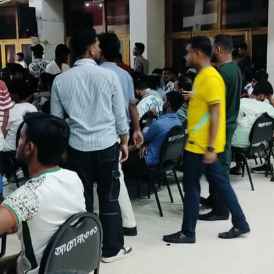 The image depicts a crowded indoor space in Lalmohan, Bangladesh, likely a community hall or meeting room. Numerous individuals, primarily young men, are present, either standing or seated on black plastic chairs. Some are dressed casually in T-shirts, while others wear button-down shirts or jerseys. A man in a yellow shirt is walking through the scene. The chairs feature text in Bengali script. The lighting is somewhat dim, and the background shows structural columns and windows. The overall atmosphere suggests a social gathering or community event.