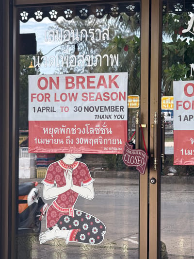 A shop in Thong Sala, Thailand, is closed for the low season, as indicated by a sign on the door written in both English and Thai. The sign reads "ON BREAK FOR LOW SEASON 1 APRIL TO 30 NOVEMBER THANK YOU" in English. Below this, the same message is written in Thai. The door also has a "COME AGAIN, WE ARE CLOSED" sign. A sticker of a traditional Thai dancer is affixed to the door, along with the shop name in Thai above the signs. The shop's dark brown door frame is visible.