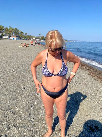 A woman in a black and white patterned bikini and dark bottoms stands on a pebble beach. She has blonde hair and is wearing sunglasses. Her right hand is on her hip and her left hand is near her bikini bottom. The sea is in the background with gentle waves. The sky is clear and blue, suggesting a sunny day. Palm trees and buildings are visible further down the beach, indicating a resort or town. Other beachgoers can be seen in the distance. The shadow of the photographer, holding a phone, is visible on the beach. The overall mood appears relaxed and summery. The time of day is likely midday due to the strong shadows. The location is identified as Marbella, Spain. There is no visible text in the image.