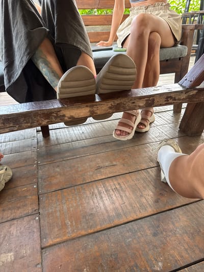 The image presents a ground-level view of several people's legs and feet in a rustic, casual setting, likely in Hà Giang 1 Ward, Vietnam. The main subjects are at least three individuals seated around what appears to be a sturdy wooden table or bench, indicated by the prominent dark wooden beam running through the center of the frame and other wooden structures.
One person, positioned towards the top left, wears light-colored shorts with a ruffled hem and light beige, chunky-soled sandals. Their bare legs are visible. Another individual, in the bottom left, wears dark, loose-fitting trousers that partially cover a distinctive blue and green tattoo on their lower leg. They are wearing light-colored, thick-soled sandals, one of which rests against a wooden support. A third person's feet are visible on the right side of the image, wearing light-colored socks and an off-white shoe or sandal, with "KEEN" faintly visible on the sole. A fourth person's arm and hand are briefly seen on the far left, reaching towards the floor.
The setting features worn, dark wooden floorboards with visible grain and small nails or screws. The overall lighting suggests it is daytime. Green foliage is visible in the background on the far left, indicating an outdoor or semi-outdoor environment, possibly a café, restaurant, or home with an open-air area. The activity appears to be a relaxed social gathering or a meal, with people seated comfortably. The rough-hewn wooden elements contribute to a natural and unpretentious ambiance.