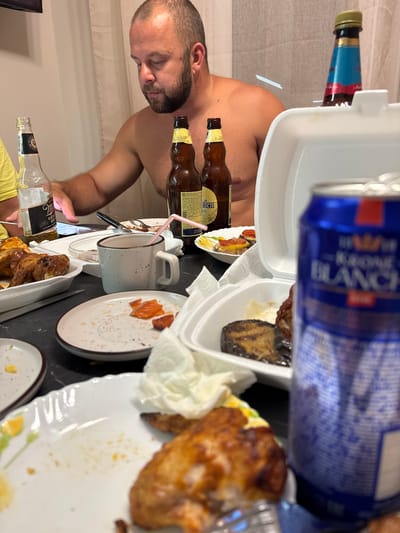 A man is sitting at a table with food and drinks. The man is shirtless and has a beard. There are several bottles of beer and a can of beer on the table, as well as plates with food. The man appears to be looking at his phone. The environment appears to be an indoor setting, possibly a home or a restaurant. The image does not provide enough information to determine the specific location in the city of Novorossiysk, Russia.