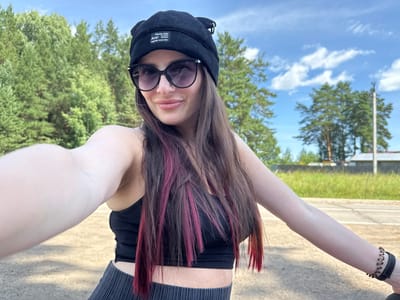 A young woman with long, dark hair with pink streaks is taking a selfie outdoors. She is wearing a black beanie, large dark sunglasses, a black ribbed crop top, and dark pleated pants. She is smiling and looking at the camera. Her right arm is extended, holding the camera. In the background, there are green trees and a blue sky with some clouds. The ground appears to be dirt or gravel, and there is a road visible in the distance. The location is identified as Rusalevka, Russia.