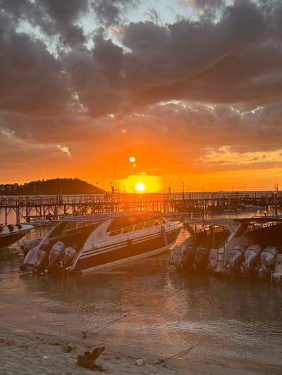 This image captures a vibrant sunset over Baan Bang Rak, Thailand. The sky is ablaze with shades of orange and yellow, illuminated by the setting sun, which appears as two distinct orbs due to atmospheric conditions. Dark, dramatic clouds drift across the sky, adding depth and contrast to the colorful display.
In the foreground, several speedboats are docked in the shallow, reflective water. These boats are equipped with multiple outboard motors, indicating their capacity for speed and transport. Their sleek designs and white and blue hulls are partially illuminated by the warm sunset light, casting shimmering reflections on the water's surface.
A wooden pier stretches out into the water, lined with lights and structures, suggesting it's a busy hub for boat activity. People can be seen walking along the pier, silhouetted against the bright sky. On the shore, a sandy beach meets the water, with a brown dog lying on the sand, looking towards the water. Further back, a green, hilly landscape with buildings can be seen in the distance, hinting at the coastal town. The overall scene evokes a peaceful yet active coastal evening.