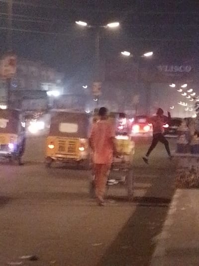 A blurry image depicts a street scene at night. Several vehicles, including a yellow tuk-tuk, are visible on the road. In the foreground, a person in an orange shirt is walking while pulling a cart. Another person is jumping or running across the street. Streetlights illuminate the area. A sign with "VLISCO" is visible in the background. Overall, the image's quality is low.