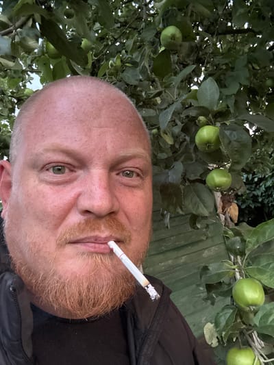 The image is a close-up selfie of a fair-skinned man with a bald head and a reddish-brown beard. He is wearing a black jacket over a black shirt and has a lit cigarette in his mouth. The man has green eyes and is looking towards the camera. He is standing outdoors, with an apple tree in the background. Several green apples are visible on the tree, along with green leaves and branches. A wooden fence is also partially visible behind the tree. The lighting suggests it is daytime, possibly late afternoon or early evening, with soft natural light. The overall setting appears to be a garden or yard. There is no specific indication of the location in the city of Лесное Снт, Russia within the image itself.