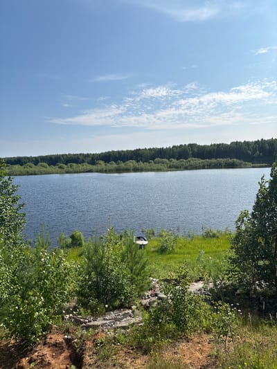 This image depicts a serene natural landscape, likely a lake or a large body of water, bordered by a dense forest on one side and a grassy embankment with some rocky outcrops on the other. The water's surface is calm, with gentle ripples indicating a light breeze. A small, white object, possibly a boat or a buoy, is visible in the water near the shore.
The sky is a clear, bright blue, dotted with scattered white, wispy clouds, suggesting a pleasant day with good weather. The sun is high in the sky, casting bright light on the scene, implying it is daytime, likely midday or early afternoon. The abundant greenery of the forest and the grassy areas signifies a season of growth, possibly summer or late spring. The presence of trees and the overall natural setting suggest a rural or wilderness area.
The perspective is from the shore, looking out over the water and across to the treeline. The overall mood conveyed is one of tranquility and natural beauty. There is no visible text in the image. The location context of Kurgan, Russia, places this scene within a specific geographical region known for its natural landscapes.