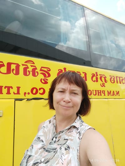 A woman with short, brown hair and light skin is standing in front of a yellow bus with red lettering. She is wearing a floral-patterned shirt. The bus has large windows that reflect the sky. The date and time "14.06.2026 12:36" are visible in the lower right corner. The setting appears to be outdoors, with a bright sky and clouds visible in the bus windows. The red lettering on the bus is in a foreign script. The location is Bavet Quarter, Cambodia.