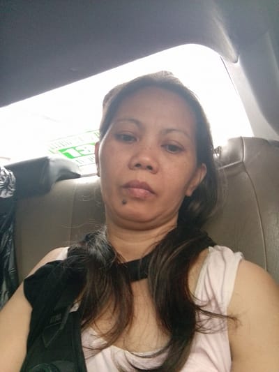 The image captures a woman taking a selfie from the back passenger seat of a vehicle, likely a car or van, in Pasay, Philippines. The woman, with long dark hair, looks directly at the camera with a neutral expression. She is wearing a light-colored top, possibly white or light pink, and has a dark garment or strap over her left shoulder. A distinct mole is visible below her lower lip.
The car's interior features a dark grey headliner above and tan or brown upholstered seats. Bright daylight streams in through the windows, indicating it is daytime. Through the rear window, a green sign or advertisement is partially visible. The most discernible text on this sign is the number "13" in green, along with some other green shapes and faint text that is too blurred to read clearly, though parts might suggest words like "LIVING" or street names. The lighting suggests a bright outdoor environment creating strong backlight for the interior shot.