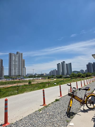 The image depicts an outdoor scene in a developing urban area, likely in Daejeon, South Korea, given the architectural style of the buildings. In the foreground, a bright yellow and brown bicycle is parked on a gravel path beside a paved walkway. The bicycle has a basket on the front and a rear rack. Orange traffic cones with reflective gray bands are spaced along the edge of the walkway, demarcating a construction or safety zone.
Beyond the walkway, there's a grassy area with some vegetation and orange construction fencing. In the mid-ground, several tall, modern apartment buildings are visible, some with distinctive gray and white facades. Further in the distance, more city buildings and some green trees are scattered across the landscape. The sky is a clear, bright blue with a few wispy white clouds. The overall impression is a sunny day in a city undergoing construction and development.