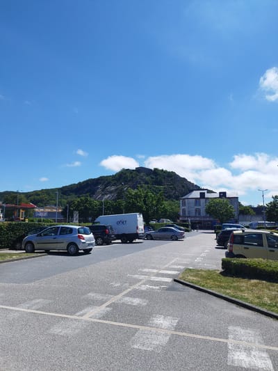 A bright, sunny day in Cherbourg-en-Cotentin, France, is depicted in this wide-angle photograph. The scene is a parking lot in front of a building with a rocky, tree-covered hill behind it. The sky is a vibrant blue with scattered white clouds.
In the foreground, a paved parking lot with white line markings and a pedestrian crossing is visible. Several cars are parked in the lot, including a silver hatchback, a dark SUV, and a yellow wagon. A white delivery van with the company name "ONET" printed on its side is also present.
Behind the parking lot, a row of buildings lines the street. One building, which appears to be a commercial establishment, has signs for "BAR TABAC KIOSQUE PRESSE". Further back, a larger, multi-story building stands out.
The hill in the background is lush with green trees, and at its summit, the ruins of a fort or castle are visible. The overall impression is of a pleasant, ordinary day in a town with a mix of modern amenities and historical features. The time of day appears to be late morning or early afternoon, given the bright sunlight and clear sky. There are no people prominently featured in the image, suggesting a focus on the environment and the objects within it.