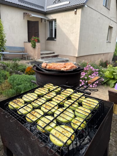 In Rezekne, Latvia, this image captures an outdoor grilling scene in a backyard. In the foreground, a black metal grill contains rows of sliced zucchini, seasoned and lightly charred from the coals below. Beyond the grill, a dark, heavy grill sits with pieces of raw meat on its surface. The backdrop features a light-colored house with a dark brown door, a small covered porch, and a few windows. Lush green foliage and pink flowers surround the house, suggesting a well-maintained garden. A grey metal bench is visible to the left, and a stone path leads towards the house's entrance. The scene implies a casual, domestic setting, possibly a family gathering or a relaxed meal outdoors. The lighting suggests it's daytime, with the sun casting soft shadows.