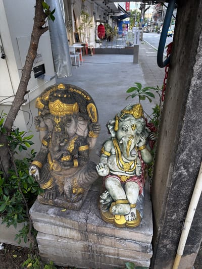 Two stone statues of Ganesha, adorned with gold accents, stand on a multi-layered concrete base next to a textured concrete pillar. The left statue is larger and darker, with multiple arms, while the right statue is smaller, lighter green, and appears to be touching its ear. Green foliage surrounds the statues. Behind them, a paved sidewalk runs alongside a street with buildings and a few vehicles. The scene is in an urban environment, likely a commercial area, as suggested by the storefronts and street activity. The style of the statues and the presence of greenery indicate a cultural or artistic element within the city. The overall atmosphere is that of an outdoor display or decoration in a public space.