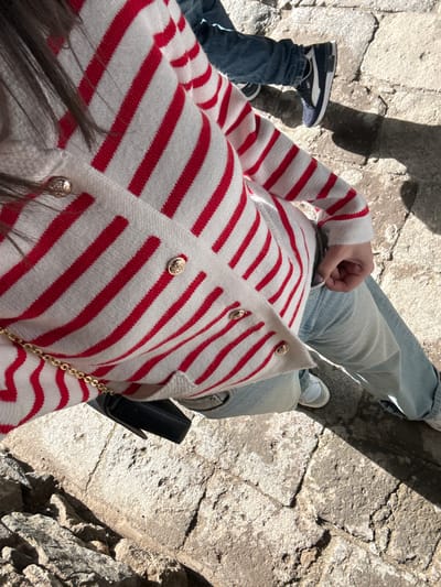 A street-level, eye-level, medium shot captures a person wearing a red and white striped cardigan, light blue jeans, and white sneakers, possibly in the city of Goght, Armenia. The person is seated on a stone surface. A small black bag with a gold chain hangs beside them. Another person, wearing jeans and black and white sneakers, is partially visible in the frame, their leg extending from the left. Strong sunlight casts a shadow of an object or person, obscuring the stone surface. The stone surface is cracked and uneven, suggesting an old or unmaintained structure.