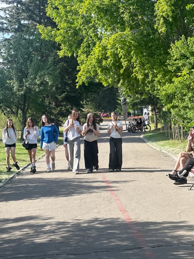 The image shows a group of six young women standing in a line on a paved path next to a grassy area and trees. The women appear to be casually dressed, with some wearing t-shirts and shorts, and others wearing long pants and athletic shoes. The time of day appears to be daytime, with sunlight casting shadows from the trees onto the path, suggesting it is a bright, sunny day. The overall mood seems relaxed and casual, as the women are standing together, possibly posing for a photo or waiting for something. There are bicycles parked in the background, indicating a park or recreational area. The women's expressions are neutral or slightly smiling. There is a red painted line on the path. The image is oriented sideways. There is no visible text in the image.