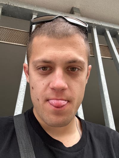 A close-up, selfie-style, eye-level shot shows a man with a buzzed haircut and sunglasses perched on his head. He is wearing a black t-shirt and a silver chain necklace. The man's tongue is sticking out, and he has a mole on his left cheek. He is positioned in front of a grey metal fence with a mesh-like grate behind it. The background is dimly lit, suggesting an outdoor or semi-enclosed space. The overall impression is casual and playful.