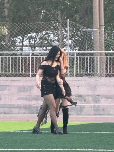 Two women wearing dark clothing are standing on a track, possibly a running track, in an outdoor setting. One woman has long black hair, and the other has long blonde hair. A chain-link fence and some trees are in the background. The location is possibly Linghe, China.