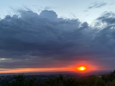 A sunset is captured in the sky above a rural landscape. The sun is a brilliant orange orb near the horizon, with rays of light extending outwards. The sky above is filled with dramatic clouds, ranging from dark gray to lighter shades of blue and white. The lower portion of the image shows a rolling landscape of green trees and fields, with hints of buildings and roads suggesting a small town or village. The overall impression is one of natural beauty and tranquility. The specific location is the Arandjelovac Municipality in Serbia. There are no people or animals visible in the image.