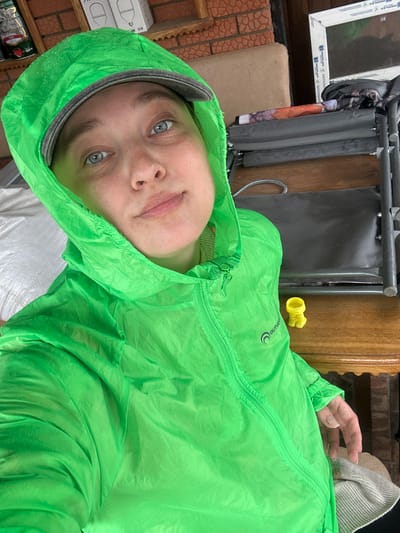 The image shows a close-up, low-angle selfie of a person wearing a bright green hooded jacket. The person's face is the primary focus, with their head tilted and eyes looking directly at the camera. They have light skin with freckles and light-colored eyes. Their lips are slightly parted, and they have a neutral expression. The jacket is shiny and appears to be water-resistant, with visible water droplets on the fabric. A zipper runs down the front of the jacket, and the hood is pulled up, framing their face. The background is blurred but suggests an indoor or semi-outdoor setting. Visible elements in the background include a wooden surface, part of a metal structure, and what appears to be brickwork. The lighting is somewhat dim, casting subtle shadows. The image is oriented vertically. There is no discernible text in the image. The activity appears to be a casual self-portrait.
