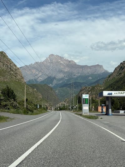 The image depicts a scenic road leading towards a towering mountain range under a cloudy sky. The road, a two-lane asphalt surface with white markings, curves to the right as it enters a valley. On the right side of the road, a gas station with a prominent blue and white sign reading "АЗС НАP" and digital price displays for fuel is visible. A white fuel dispenser is present. Power lines stretch across the sky, supported by tall metal towers. The mountains flanking the road are covered in lush green vegetation and rocky outcrops. The overall scene is one of a natural landscape with man-made infrastructure present. The location is the city of Mizur, Russia.