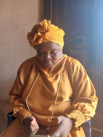 An adult female is depicted seated indoors in Ota, Nigeria. She has a darker complexion and is wearing a gold-colored ensemble comprising a headwrap with a prominent ruffled embellishment and a long-sleeved garment. The garment features a hooded neckline with a drawstring and elasticized, ruched cuffs. Silver-colored, patterned earrings are visible on both ears. Her head is bowed, and her gaze is directed downward towards a dark-colored mobile phone, which she holds horizontally with both hands. She is seated on a chair with visible dark blue plastic components. The immediate background consists of a plain light brown wall on the left and a darker, possibly wooden or painted surface on the right.