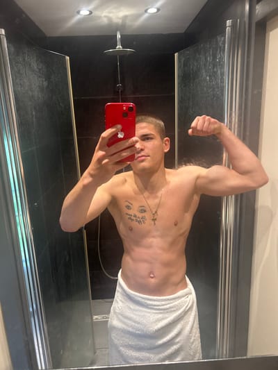 A man stands in a shower in Ede, Netherlands, holding a red smartphone while posing for a mirror selfie. He is shirtless and partially wrapped in a white towel. He is flexing his right bicep and has a gold chain with a cross pendant around his neck, as well as tattoos on his upper chest. The shower has black tiled walls and a silver showerhead. The mirror's reflection shows the man holding the phone with both hands.