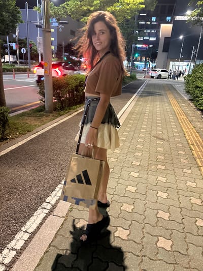 A young woman with long brown hair smiles at the camera while holding two shopping bags, one with the Adidas logo. She is wearing a brown cropped jacket over a white and grey layered dress, and black shoes. She is standing on a paved sidewalk next to a street. The scene appears to be in an urban area with buildings and streetlights visible in the background. The time of day is likely evening or night, as indicated by the artificial lighting and shadows. The weather is not discernible from the image. The text "Adidas" is visible on one of the shopping bags.