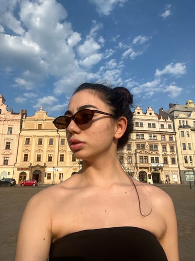The image shows a young woman with dark hair pulled up in a bun, wearing sunglasses and a black strapless top. She is looking to her right, with her shoulders and upper chest visible. In the background, there are several buildings with ornate facades and windows, suggesting an urban European setting. A few cars are parked in the street in front of the buildings, and a cobblestone plaza is visible. The sky is blue with scattered white clouds. The woman's facial features, including her lips and cheekbones, are prominent. The lighting suggests it is daytime, possibly late afternoon due to the long shadows. The context suggests the location is in the city of Pilsen, Czechia, given the architectural style of the buildings and the visible signage which appears to be in Czech. Specifically, the building with the "ANTIQUE" sign and the text above it could be in the historic center of Pilsen, a common tourist area.