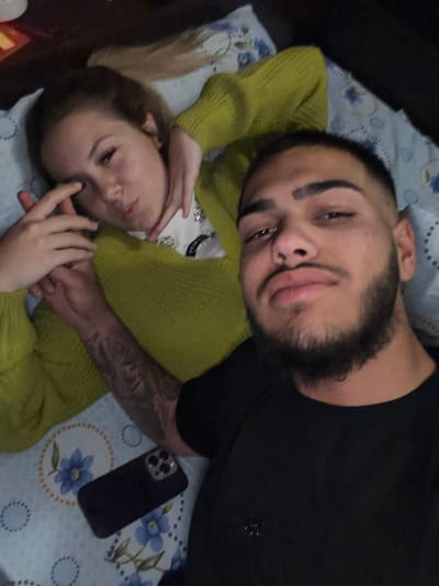 A man and a woman are lying down on a surface patterned with circles and blue flowers. The man, on top, has dark hair, a beard, and a piercing in his nose. He is wearing a black shirt. The woman below him has light hair and is wearing a lime green sweater over a white top with embellishments. The man's tattooed arm is visible, and a black smartphone is present on the patterned surface. No location context of Radomir, Bulgaria, can be confirmed from the image.