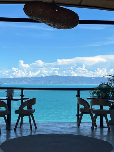 This is a vertically oriented, medium shot of a balcony overlooking a body of water. The scene suggests a tropical location, possibly a resort. In the foreground, on the left, is a large, woven decorative object, possibly a lamp or sculpture, hanging from above. On the right side of the frame, two minimalist wooden chairs are visible, one behind the other. The chairs are positioned on a floor that appears to be made of light-colored wood or tiles, with a circular rug underneath.

Beyond the balcony, a vast expanse of turquoise water stretches to the horizon. The water is calm, reflecting the bright blue sky. A distant coastline, dotted with vegetation and possibly buildings, is faintly visible across the water. A scattering of white, cumulus clouds are present in the sky, suggesting fair weather. The overall mood is serene and relaxed, evoking a sense of vacation or leisure. The lighting indicates it is daytime, likely mid-morning or early afternoon, given the bright sunshine and clear sky. No people are visible in the image, and there is no visible text. The orientation of the image is rotated 90 degrees clockwise, so the scene is viewed sideways.