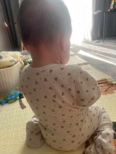 The image shows a baby from behind, sitting on a yellow playmat. The baby is wearing a white onesie with a pattern of small pink hearts and black dots. To the left of the baby, there is a white woven basket filled with light-colored stuffed toys and a blue toy fish with yellow fins and spots. In the background, sunlight streams through a sheer curtain, casting striped shadows on the floor and playmat. A dark furniture item is visible on the right. The scene appears to be indoors, likely in a living room or playroom. There is no information available to indicate the location in the city of Voronezh, Russia.
