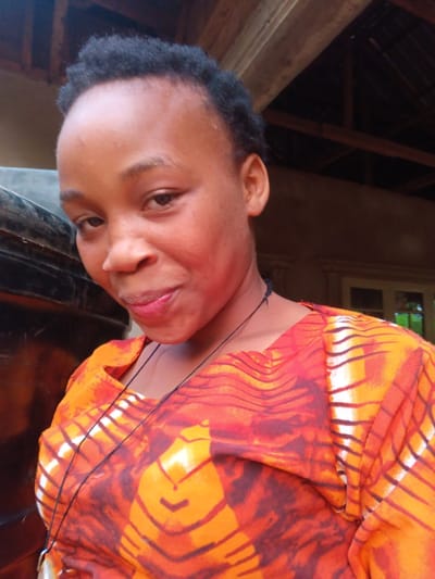 A woman with short, dark hair and light skin is centered in the frame. She wears a vibrant orange and white patterned top. A thin black cord, possibly a necklace, is visible around her neck. To the left, a dark, rounded object is partially visible. The background suggests an outdoor or semi-outdoor setting, with a building structure featuring visible beams and a window. The overall lighting is soft and natural.