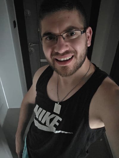 A man with dark hair and a beard is smiling and wearing glasses and a black Nike tank top with a silver chain. He appears to be indoors. The location is unknown.