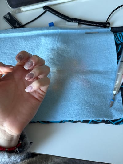 The scene, located in Hull, United Kingdom, displays a close-up, elevated perspective of a human left hand positioned on a light blue disposable mat. The fingernails of the index, middle, ring, and pinky fingers show partially removed nail product, exposing areas of the natural nail plate. Brownish-red discoloration is visible beneath the middle and ring fingernails. The mat is soiled with fine, pale pinkish-white dust, consistent with nail filing debris. To the right of the mat, a silver electric nail drill, fitted with a blue-collared bit, is positioned vertically. In the upper left background, portions of a white electronic device, a white stylus, and a black coiled cable are visible, alongside a small blue plastic object. A black fabric with blue geometric patterns is visible beneath the disposable mat. The individual's wrist features a red beaded bracelet, and a grey, textured garment is partially observed at the bottom left. The objects are situated on a white, textured surface.