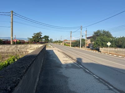 The image depicts a street scene in Shishmantsi, Bulgaria, under a clear blue sky. A paved road runs through the center of the frame, with a sidewalk on the left side separated by a low concrete wall. Utility poles with numerous wires are visible along the road, with a concentration on the left side. Houses and trees line the right side of the road. A stop sign is visible on the left, near a utility pole. In the distance, mountains are faintly visible. The overall impression is a quiet, semi-rural street during daylight hours.