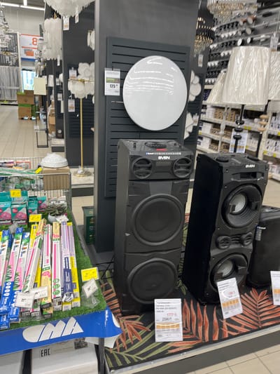 The image shows a retail display of audio equipment and lighting fixtures inside a store in Бараўлянскі сельскі Савет, Belarus. In the foreground, there are two large black speakers, branded "SVEN," stacked vertically. The right speaker is a tall, narrow unit with visible woofers, while the left speaker is a wider, shorter unit. Between the speakers and extending to the left, there is a display of fluorescent light bulbs in various packaging, some of which have price tags. A metallic cart filled with more light bulbs and other items is visible to the left. In the background, shelves are stocked with more lighting products, including chandeliers and lamps. A large, circular white light fixture is mounted on a dark wall display to the left of the speakers. A price tag for the circular light is visible, showing "125." The overall environment suggests a large retail store specializing in home goods and electronics. The flooring under the speakers has a tropical leaf pattern.
