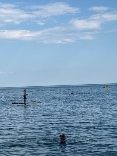 This image depicts a serene view of the Black Sea in Partenit, Ukraine, under a partly cloudy sky. In the foreground, a woman is swimming. Further out, a man stands on a paddleboard, holding a paddle. In the distance, several other people are visible in the water, and a small yellow boat is moving across the sea. The water is calm, with gentle ripples reflecting the sky. The overall scene suggests a leisurely summer day at the beach.