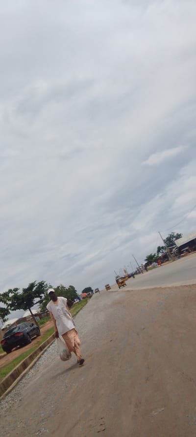 A person walks along a dusty road, carrying a plastic bag filled with items. They are dressed in a light-colored tunic and pants, with a white cap on their head. The person is walking away from the camera, with their back mostly turned. The road is unpaved and appears to be under construction, with loose gravel and dirt.
In the background, there are several vehicles, including cars and what appear to be auto-rickshaws or tuk-tuks, suggesting a bustling environment. Trees line the side of the road, and in the distance, there are buildings and structures. The sky is overcast with scattered clouds, indicating a possibly humid or rainy day. The lighting suggests it is daytime, likely mid-morning or afternoon. The scene appears to be a street in Suleja, Nigeria, given the presence of typical transportation and architecture found in the region. No specific text is legible in the image. The overall mood is neutral, depicting a common street scene in a developing urban or semi-urban area.