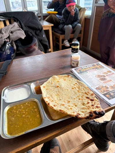 In Nyoma, India, a metal thali dish rests on a wooden table in front of a menu titled "DOLKER PUNJAB DHABA." The thali contains a portion of chickpea curry, a small container of white yogurt, and a large, golden-brown flatbread, likely naan, which is slightly charred on the edges. Beside the thali, two small containers, one black and gold and the other white with a label, are also on the table. In the background, a young person wearing a pink cap and a dark jacket sits on a wooden chair. To the left, a dark jacket and a blue helmet are visible on a chair. Further in the background, through a window, a glimpse of the outside environment can be seen, suggesting a setting with natural light and potentially a mountainous landscape common to Nyoma.