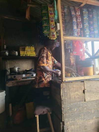 In Madalla, Nigeria, a person stands behind a wooden counter in a small shop. The shop is filled with various items, including food products displayed in plastic packaging hanging from the ceiling. There are also pots and pans on a stove, suggesting cooking is taking place or has recently occurred. The environment appears rustic, with wooden shelves and walls. The person is wearing a brightly colored shirt and is holding what appears to be a cup, possibly in the process of serving a customer or preparing a drink. The shop is dimly lit, with some natural light filtering in from an opening.