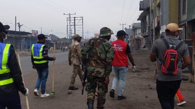 The image captures six males walking away from the camera on a dirt and gravel street in Rubavu, Rwanda, under an overcast sky. From left to right, a partially visible individual wears a black top, a yellow-green and blue reflective vest, and white gloves. Next, a male wears a black patterned shirt, blue jeans, white sneakers, a blue and yellow-green reflective vest labeled "SERVICE URBAIN," and carries a broom with a green head. Following him is a male in a tan camouflage military uniform. In the center, a male in a green camouflage military uniform and boonie hat carries a rifle slung over his shoulder. Next, a male wears a red t-shirt with a white logo and text including "GOMA," light blue jeans, dark shoes, and white gloves. The final male, partially visible on the far right, wears a grey polo shirt, black pants, a tan baseball cap, and a red backpack, and carries a wooden stick. The street surface is unpaved in the foreground, with paved sections visible further down. The immediate environment includes multi-story commercial and residential buildings lining the street, utility poles with overhead lines, and a large, empty billboard structure.