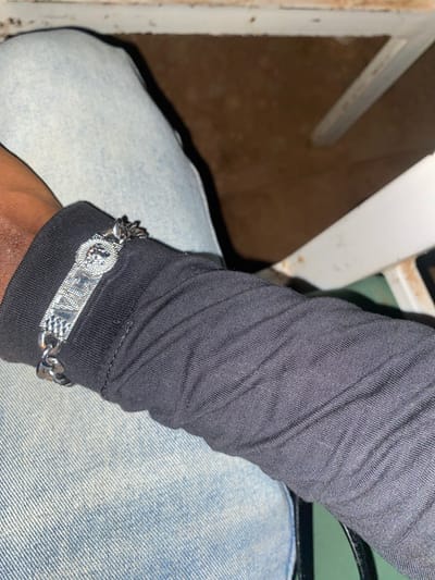 In the city of Wumat, Nigeria, a close-up shot captures a person's arm wearing a black long-sleeved shirt and light blue jeans. A silver chain bracelet with a Medusa head emblem and intricate detailing is visible on the wrist. The background is blurred, showing a brown textured surface and parts of a white chair.