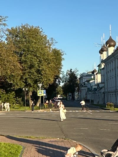 The image shows a street scene in Yazvitsevo, Russia, under a clear blue sky. The foreground features a pedestrian walkway with brick paving and a strip of green grass. A baby is visible lying on the ground, with a stroller to the right. The street itself has visible lane markings.
In the middle ground, several people are present. A woman in a long, light-colored dress is walking across the street, holding a handbag. Another person, wearing white, is on a bicycle on the opposite side of the street. Further back, more pedestrians are visible near a street sign and lampposts. To the left, a group of people are standing near a building and trees.
The background of the image is dominated by a large building complex with distinctive onion domes, characteristic of Russian Orthodox architecture. The building is white with light-colored trim and multiple windows. Trees, some with autumnal colors, line the street, and more buildings are visible in the distance. The overall impression is a sunny day in a historic urban environment.